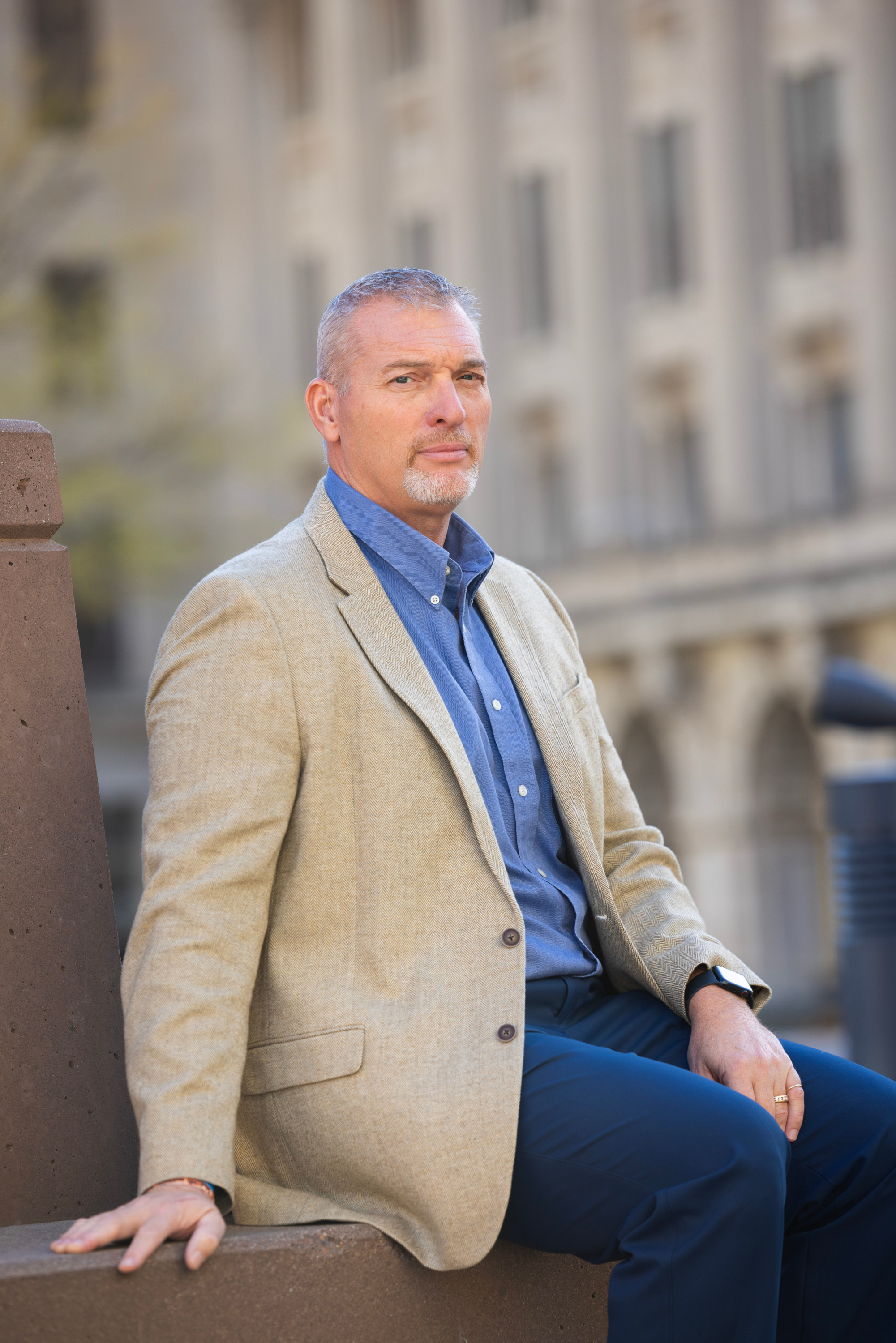 A man with gray hair and a gray goatee wearing a brown blazer, blue dress shirt and navy pants sits on a stone bench with an out-of-focus building in the background.