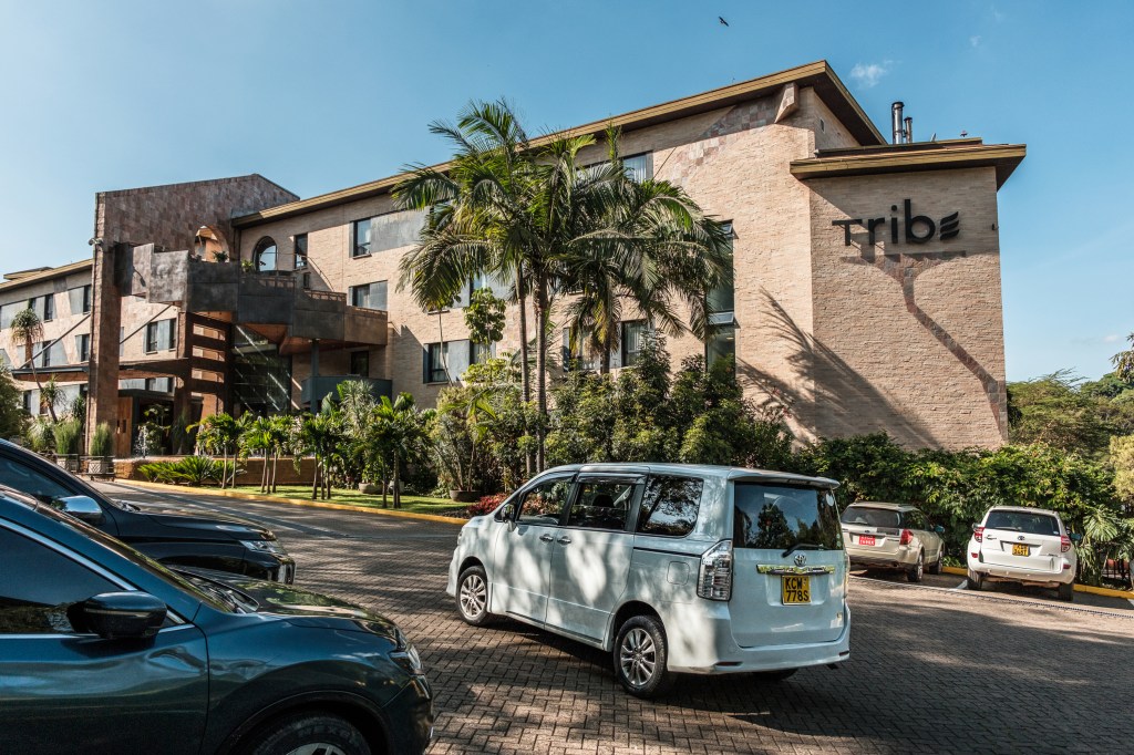 A large, luxurious-looking hotel with an ornate entryway, palm trees and the word “Tribe” written on the side of the building, seen from the parking lot as a van passes by.