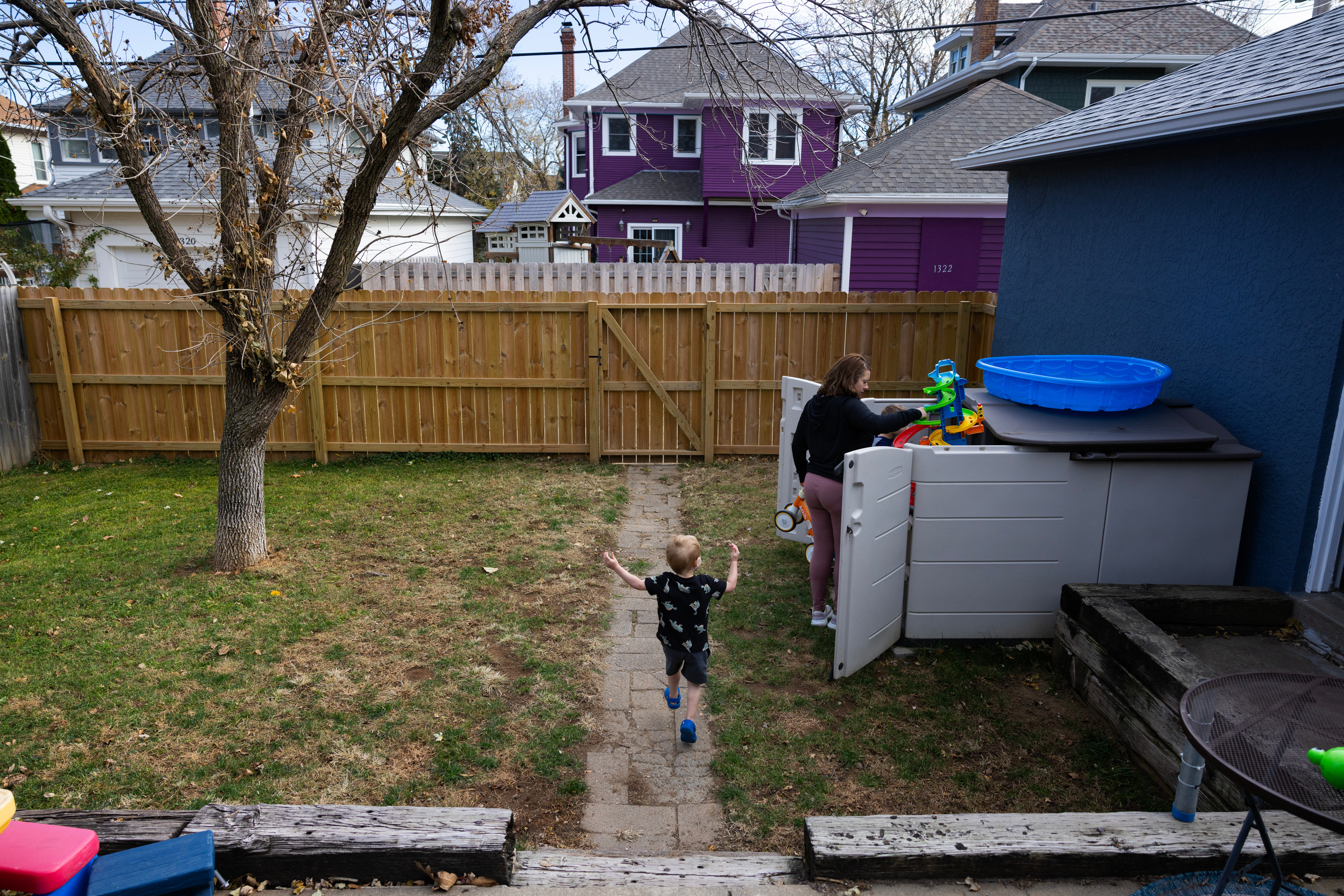 In a backyard, a small blond boy runs down a narrow stone path to a woman putting away toys in a shed. The yard is fenced in and looks out onto other two-story houses.