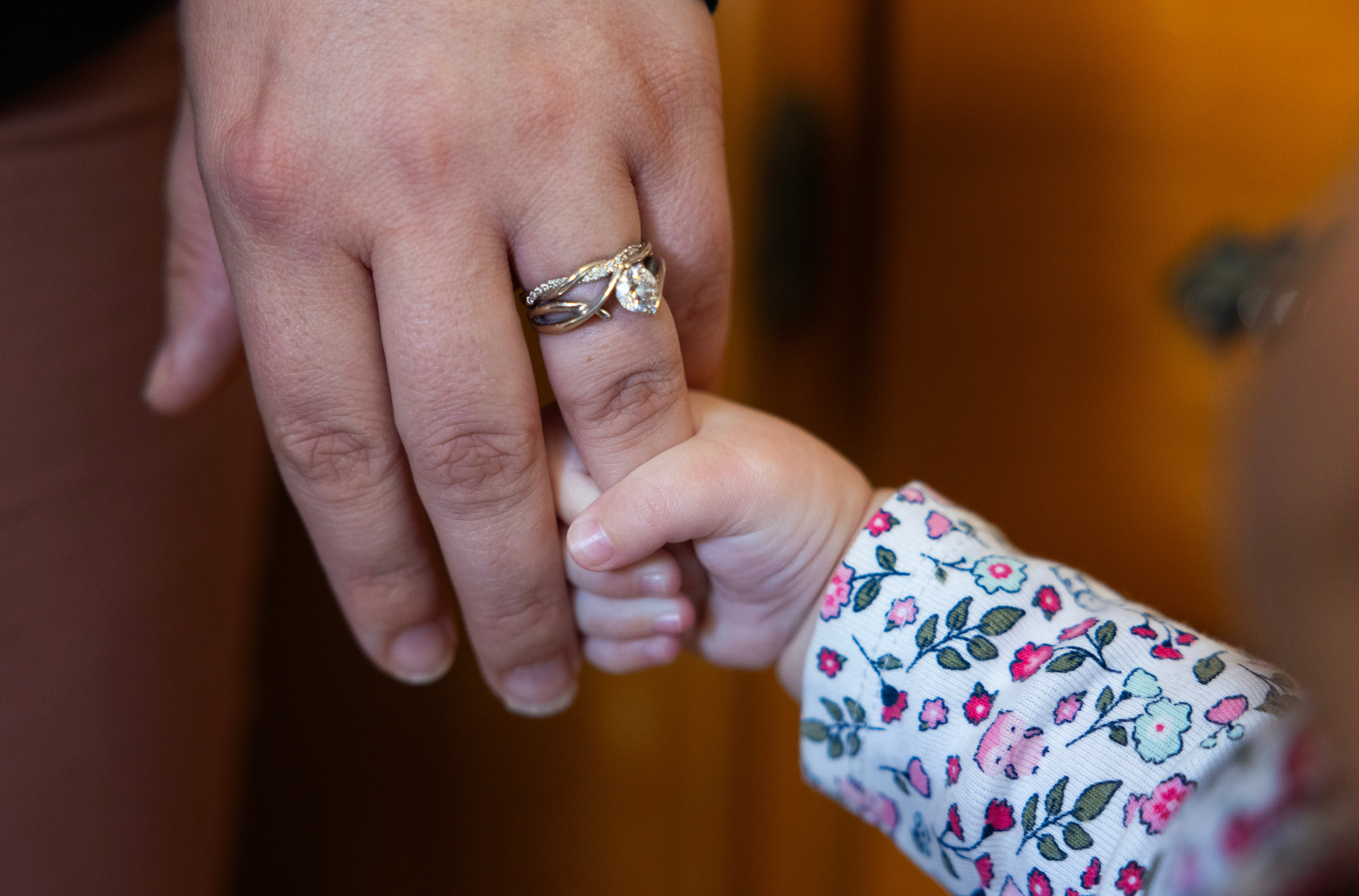 A baby’s hand grips the ring finger of an adult’s hand, which is adorned by a braided metallic ring.