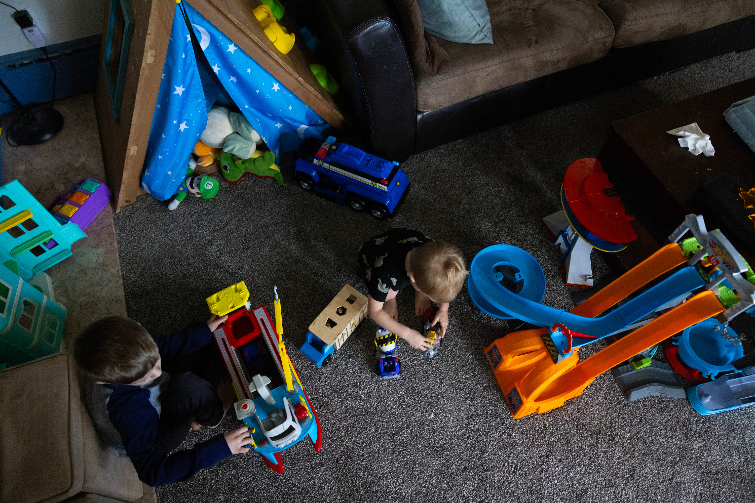 An aerial view of two children playing with brightly colored toys on a carpet between couches. A tent with stuffed animals is behind them.