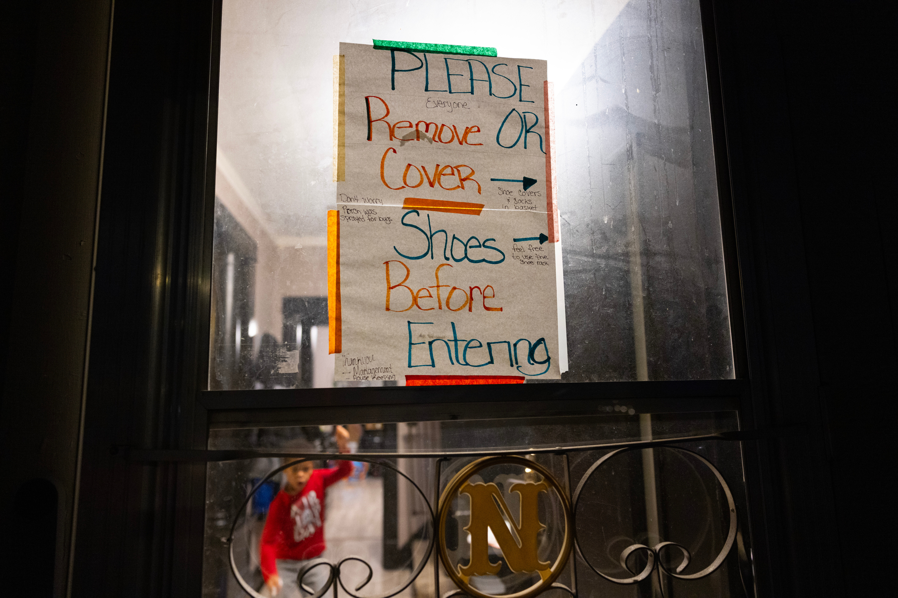 A sign reading “Please remove or cover shoes before entering” is taped to a door. In the background a small child wearing a red sweatshirt plays on the floor.
