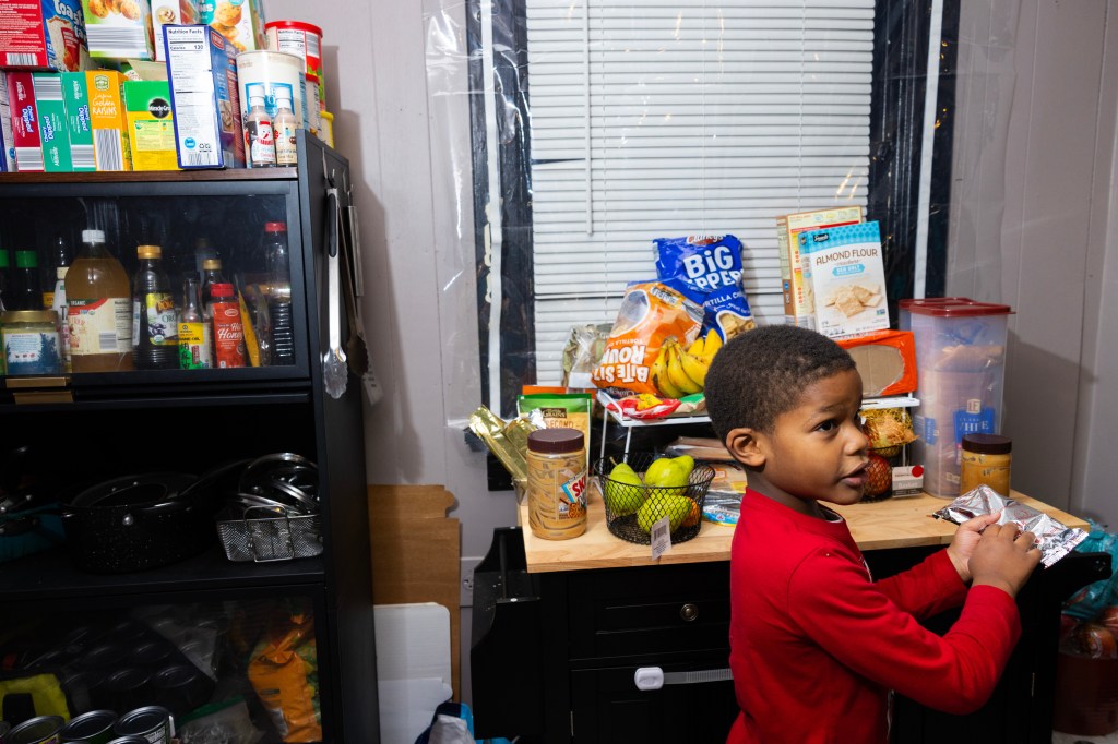 A small boy in a red long-sleeved shirt holds a metallic package in his hands in a room with different packaged food and fruit on shelves. The window has closed blinds and is covered in clear plastic.