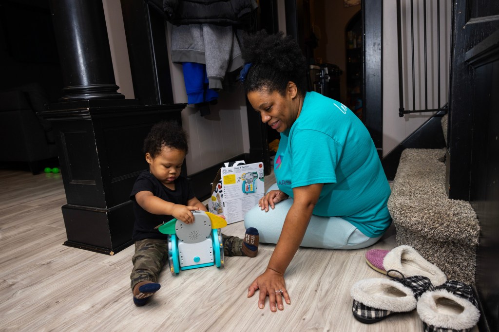 A woman wearing a teal shirt and pants and her hair in a bun sits with a baby with black curly hair on a wooden floor next to carpeted steps.