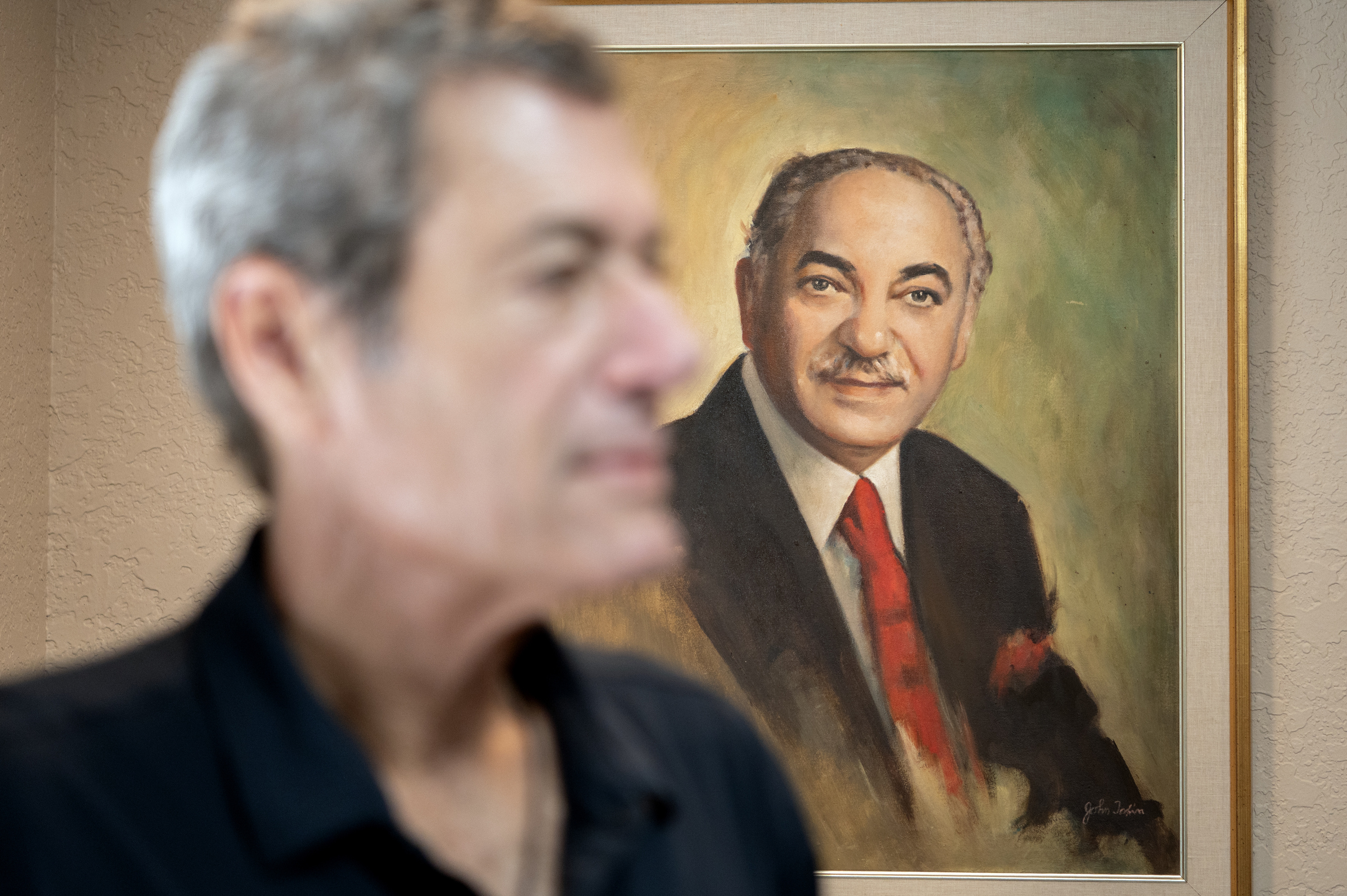 A painting of a man with a moustache and grey hair wearing a suit and tie is in focus. Jon Esformes’ profile is in front of the painting, not in focus.