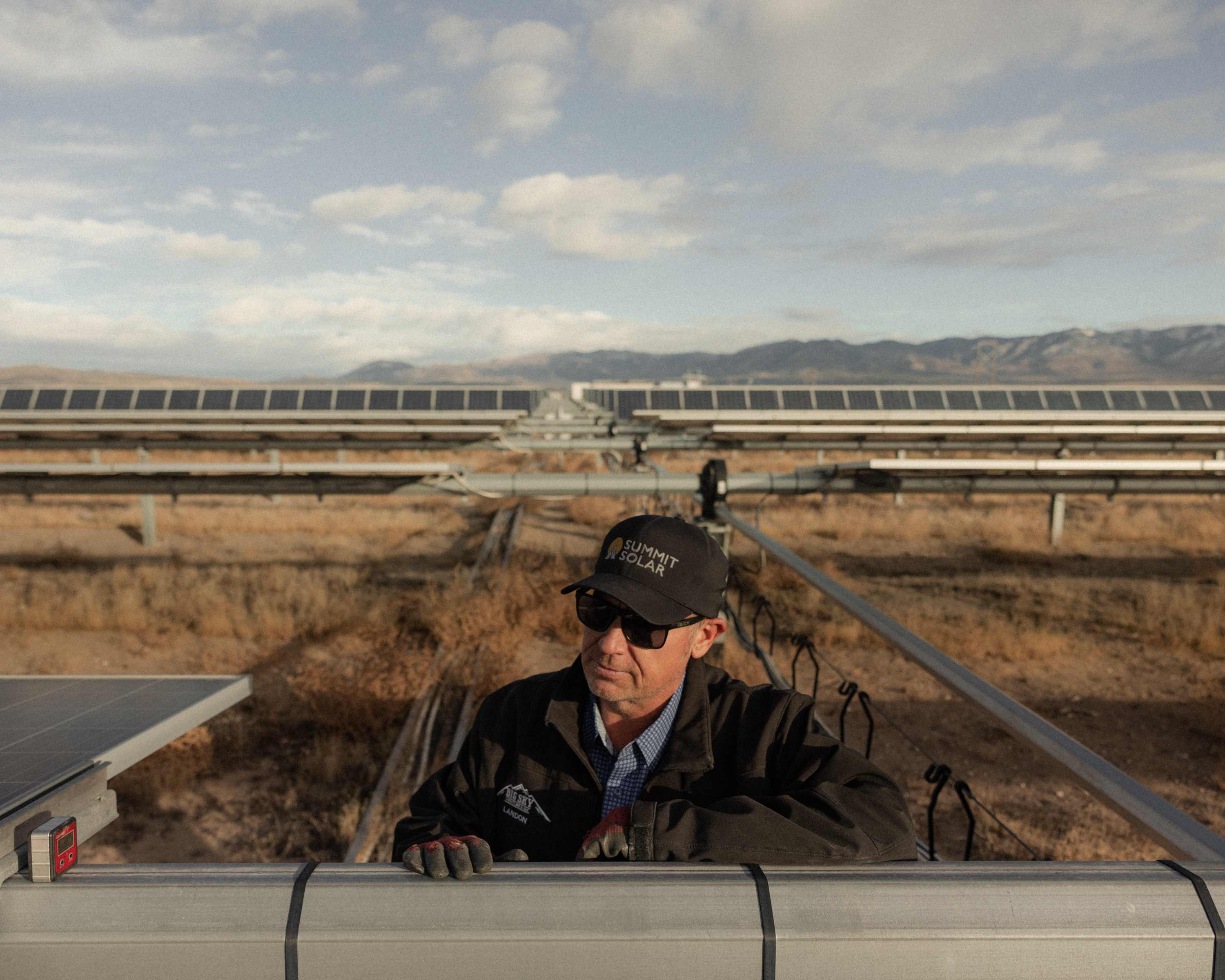A man wearing a baseball hat, sunglasses, a jacket and work gloves leans against the support bar for a solar panel array in a large field with solar panels and mountains behind him.