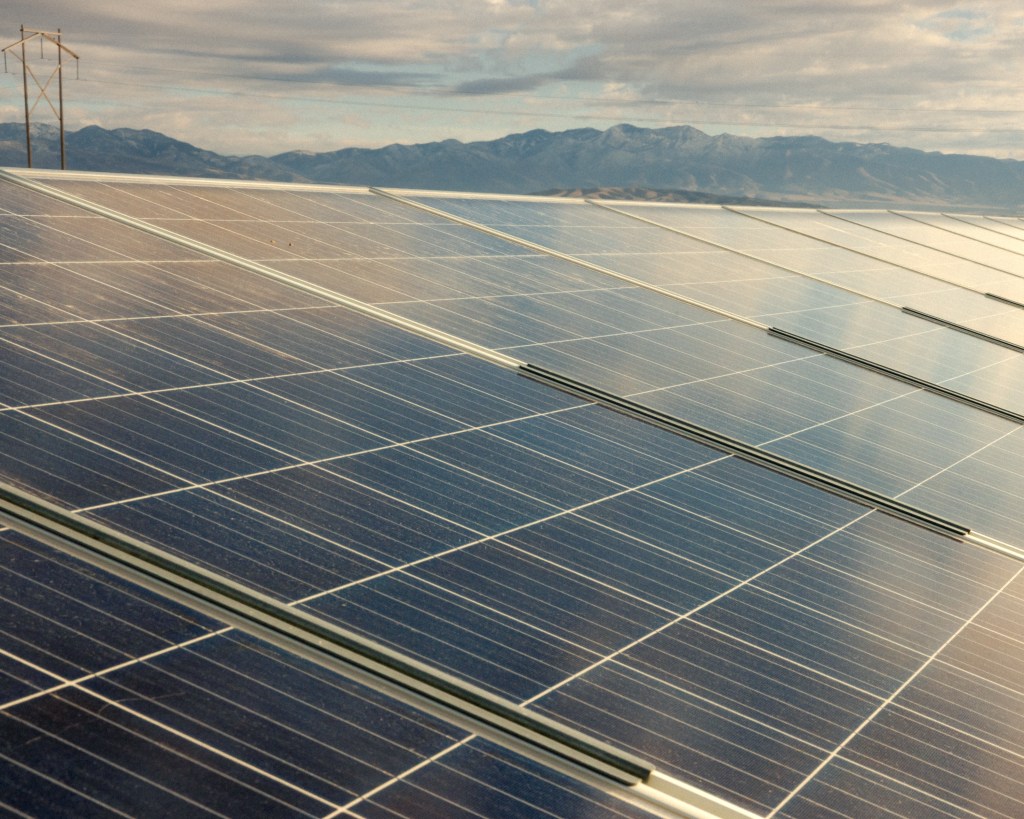 A row of solar panels under a cloudy sky, with mountains in the background.