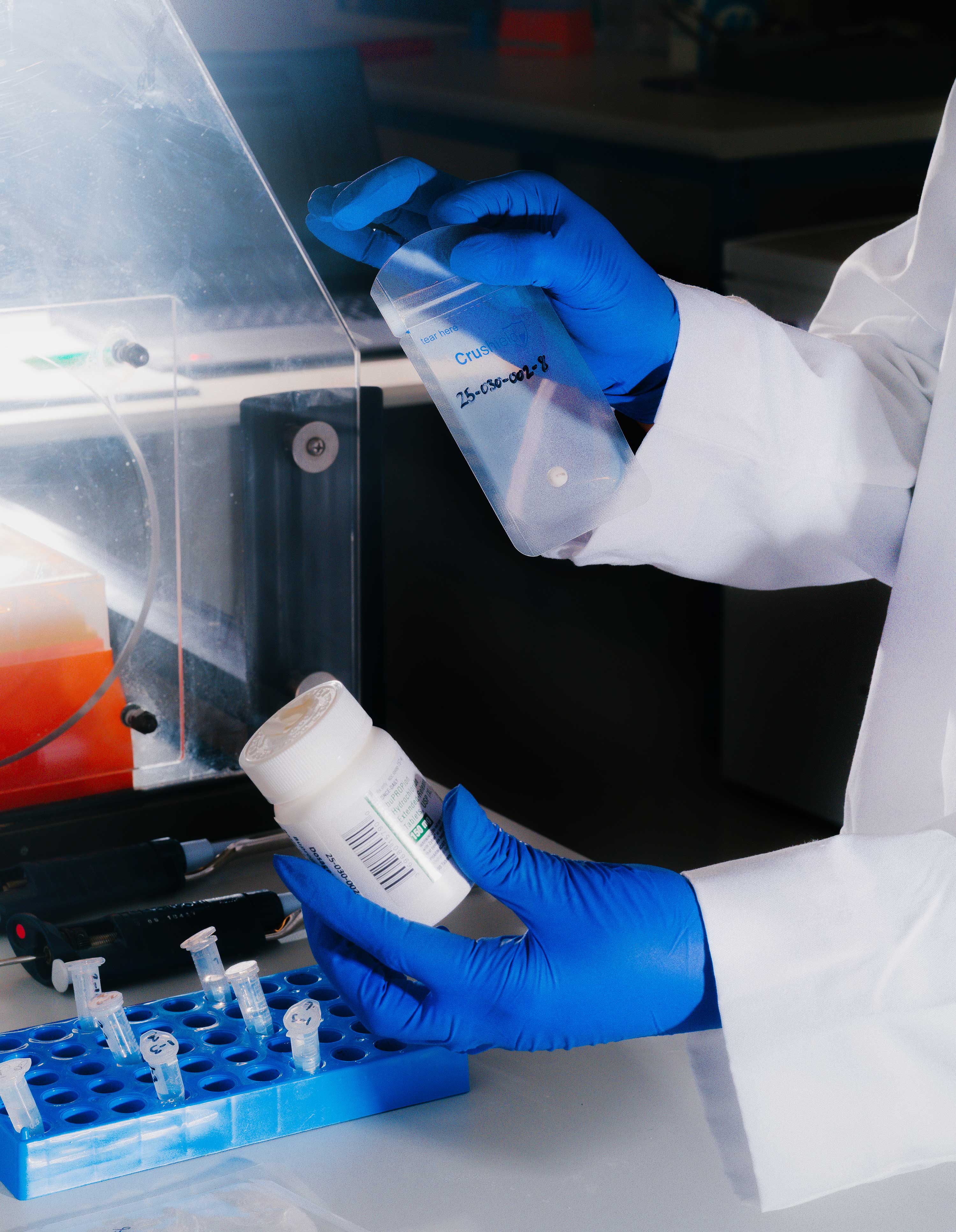 A lab technician holds a prescription bottle in one hand and a pill test sample in a plastic bag in the other.