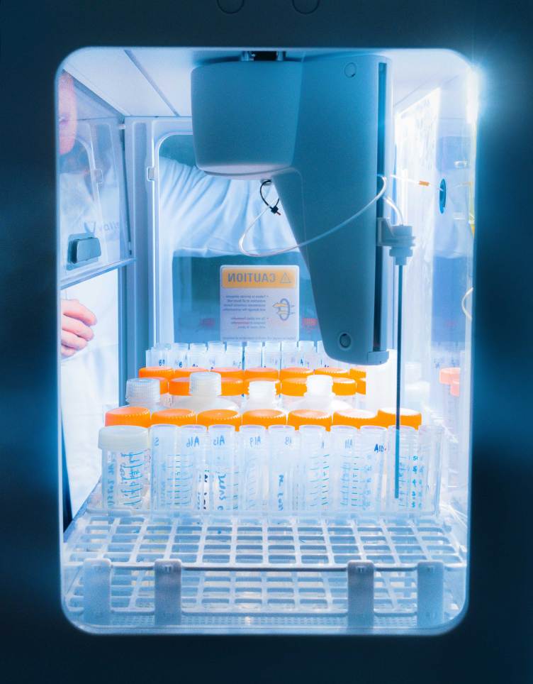 A rack of test tubes in a sealed, brightly lit box where a white robotic arm is inserting a thin piece of metal into one of the tubes.