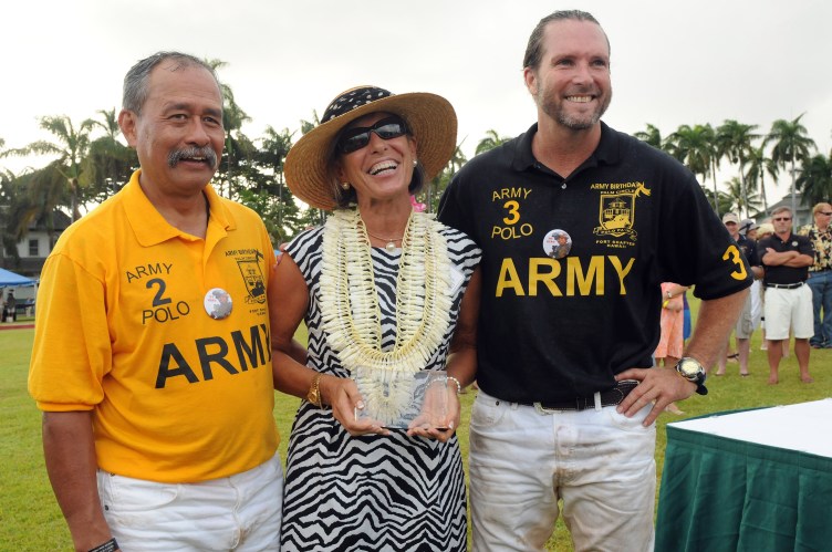 Une femme et deux hommes se tiennent dehors et sourient à la caméra. La femme tient un trophée et les hommes portent tous deux des chemises sur lesquelles sont inscrits « Armée » et « Polo ».
