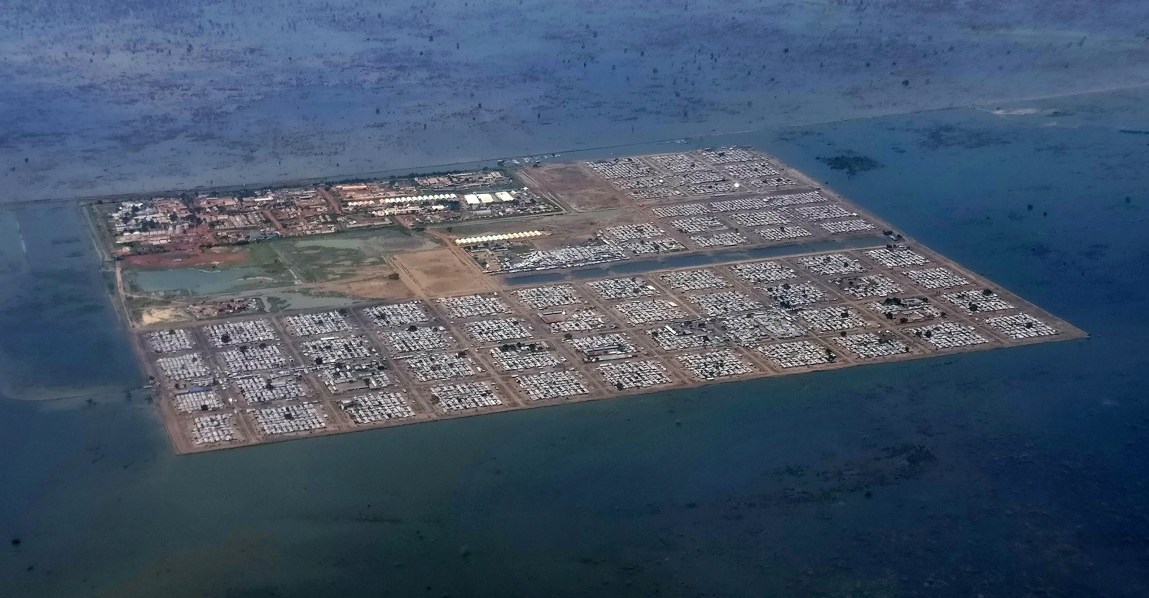 A view from above a square patch of land in the midst of blue water. The land is divided by dirt paths into square areas containing the roofs of thousands of small shack structures.