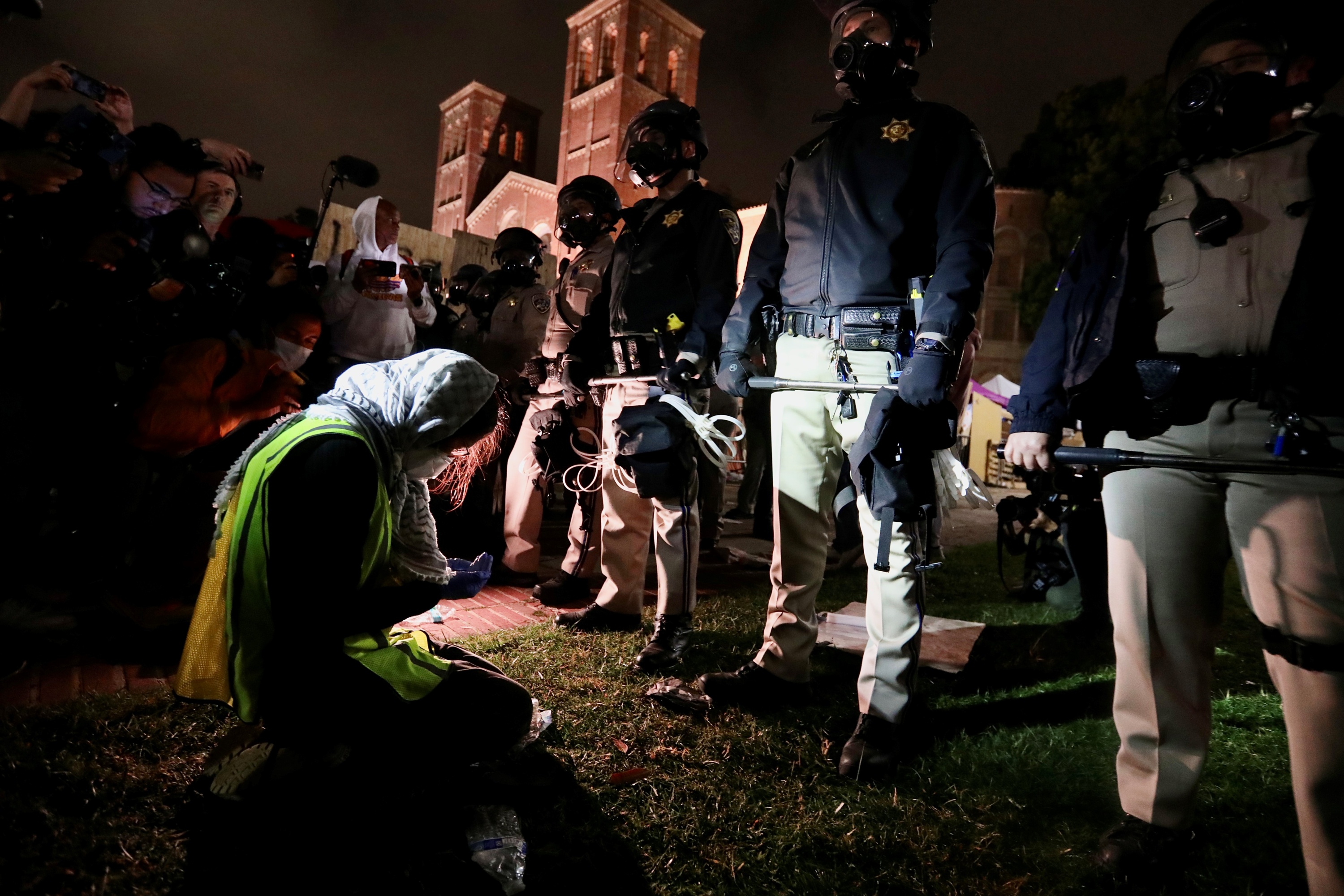 Police officers wearing gas masks and holding zip ties and metal bars stand in a line in front of a kneeling person wearing a face mask and a keffiyeh on their head. People with microphones, phones and cameras stand to the side.