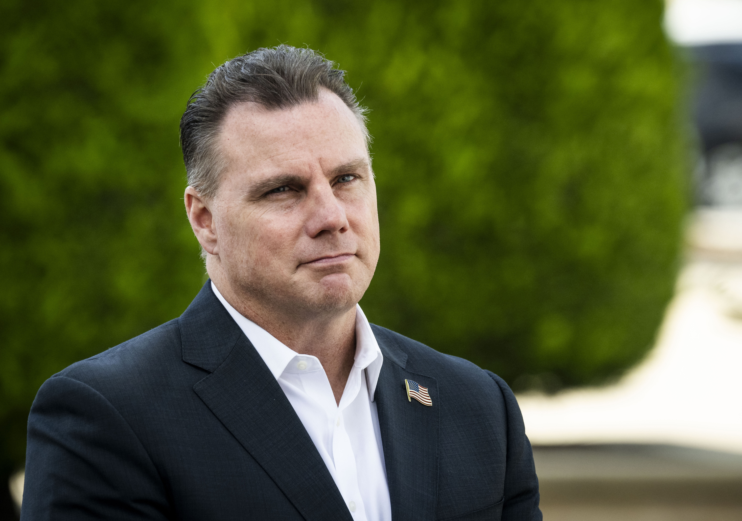 A man with gray and dark brown hair stands in front of a green bush wearing a navy suit with a U.S. flag lapel pin.