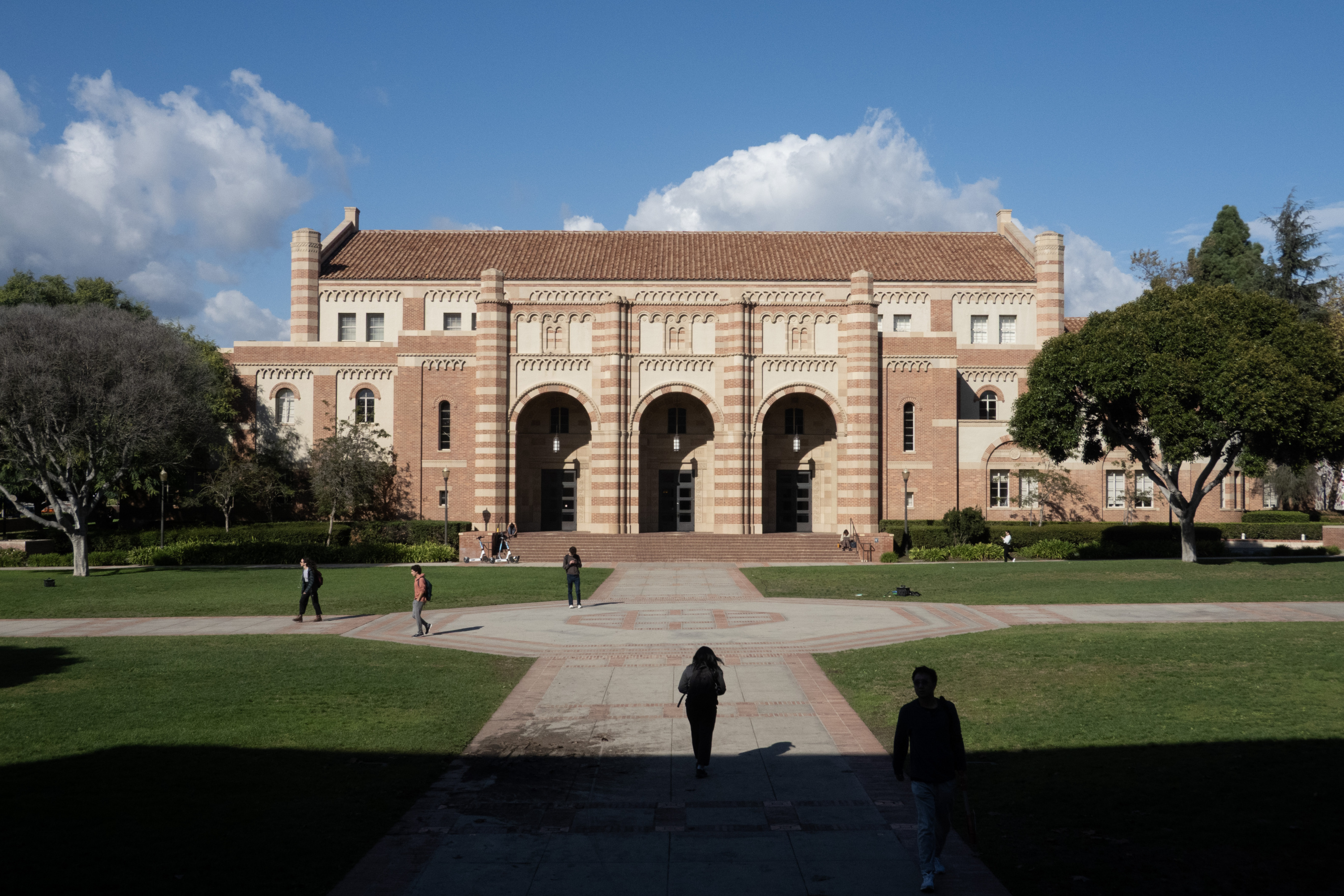 A brick building with three vaulted entryways sits at the end of a path on a college quad with green grass, trees and blue sky. A dark shadow looms at the bottom of the photo.