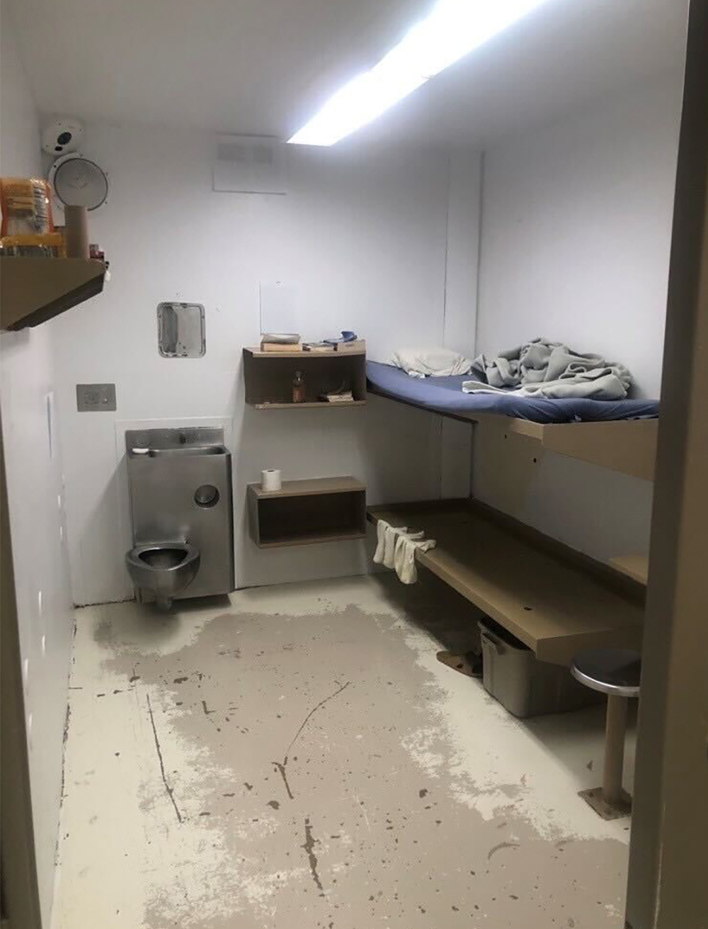 Jail cell with a metal toilet, two shelves and two beds, one above the other, with a thin pad on the upper one. The painted floor is scuffed and the top layer of paint is worn down in the central area of the cell, and bright fluorescent lights starkly illuminate the room.