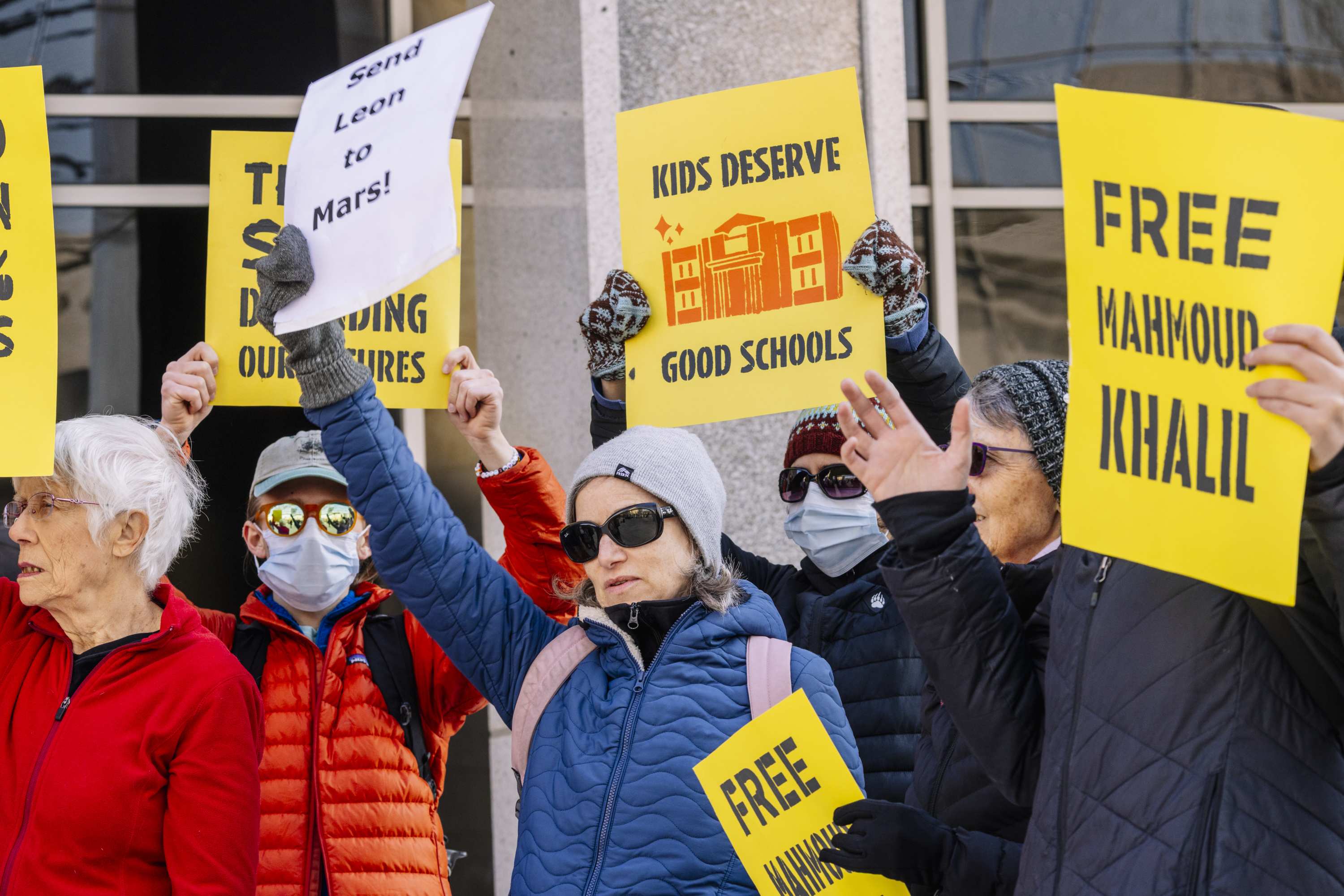 A group of people, some wearing masks and sunglasses, hold signs during a protest in front of the Department of Education building in Washington, D.C.