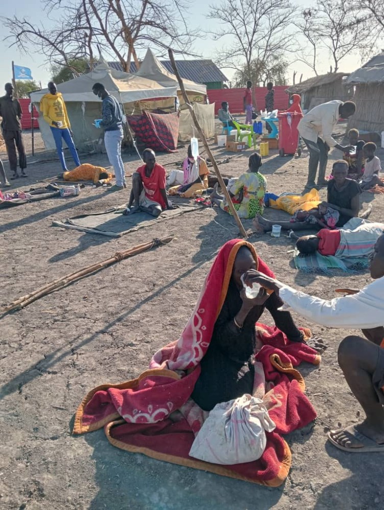 A person sits on the ground outdoors. Another person lifts a cup of water to their mouth. In the background, people sit on the ground, and other people stand over them, some distributing water. IV bags hang from sticks.