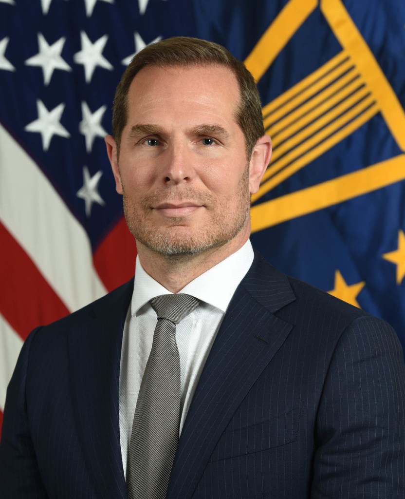 A headshot of a man in a suit looking at the camera, in front of the American flag.