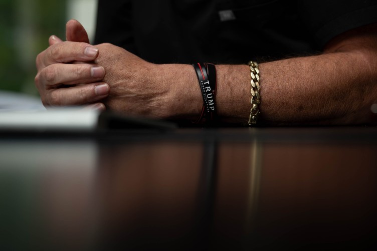 A man’s hands and arm rest on a table. He has a gold bracelet and a rubber bracelet that reads “Trump 2024.”