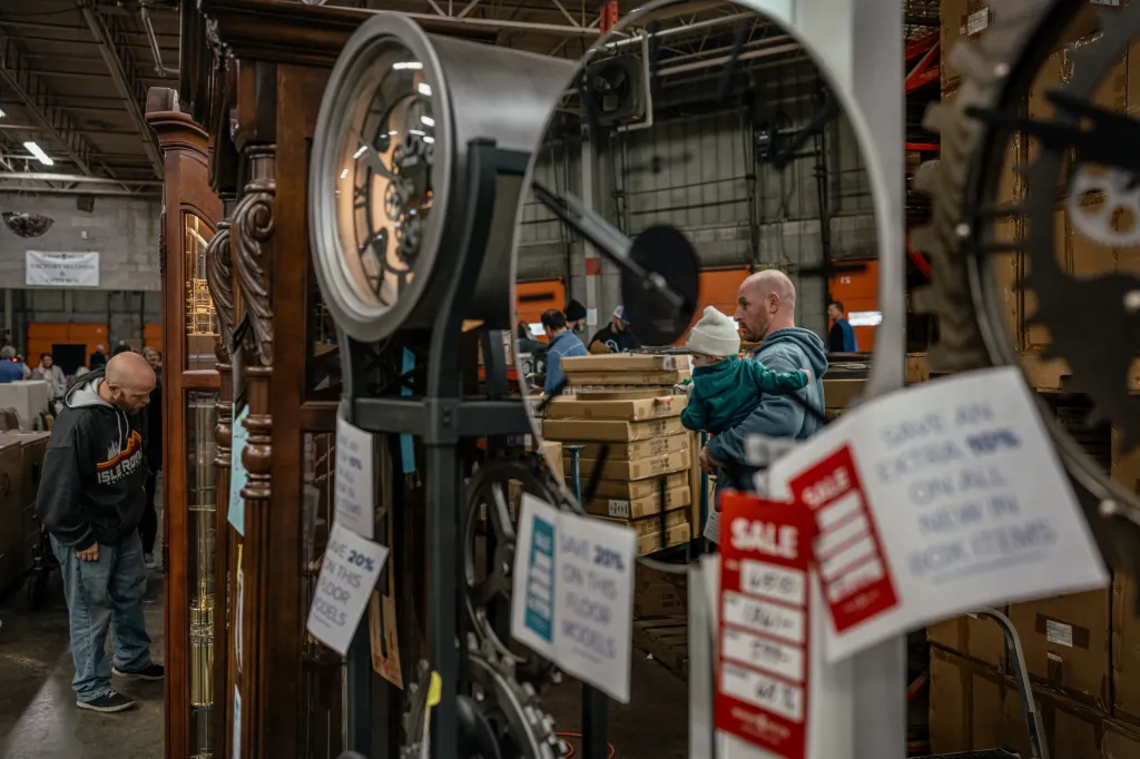 One man leans down to closely examine a grandfather clock, while another man is reflected in the mirrored face of another nearby clock.