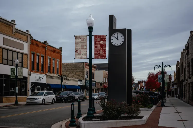 A small downtown area with cars parked along both sides of a two-lane street, storefronts and a large clock along the sidewalk.