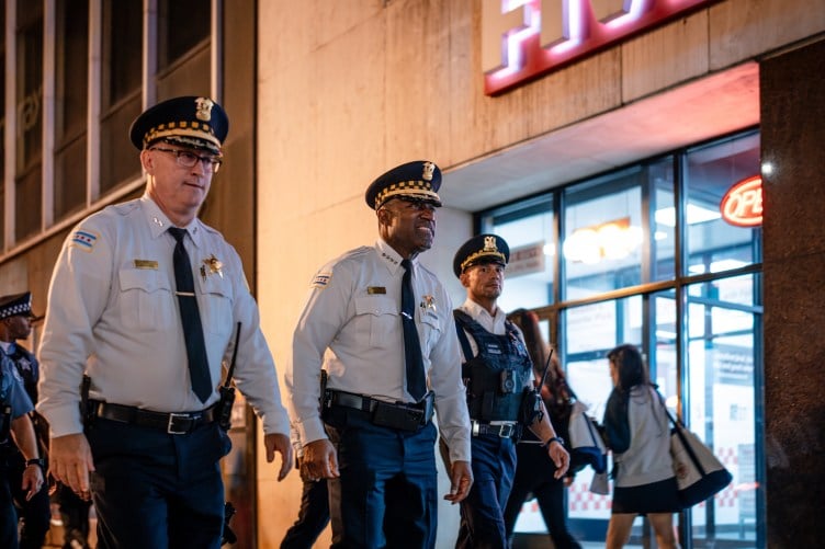 Three uniformed Chicago police officers walking along a city street at night, with two women passing in the background.