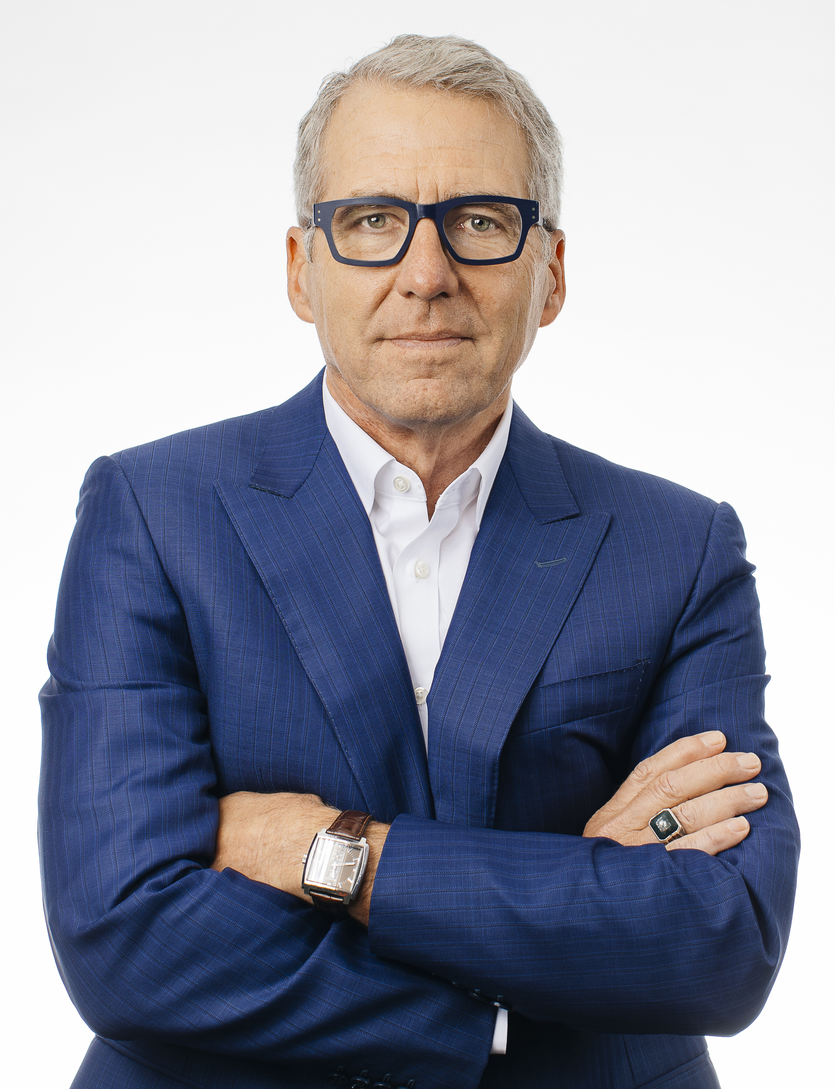 A man with gray hair wearing a blue suit and glasses crosses his arms and looks directly at the camera in front of a solid gray background.