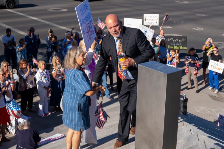 A man in a suit and tie stands tearfully at the top of concrete courthouse steps, holding the arm of a woman standing next to him. Behind him, a crowd of people applaud and hold up protest signs.