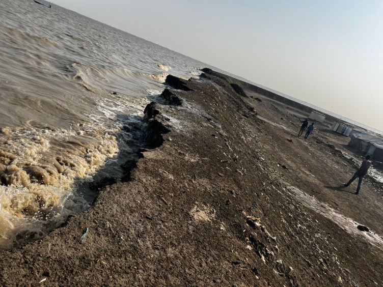 Waves crash against a collapsed embankment as people walk by in the background.
