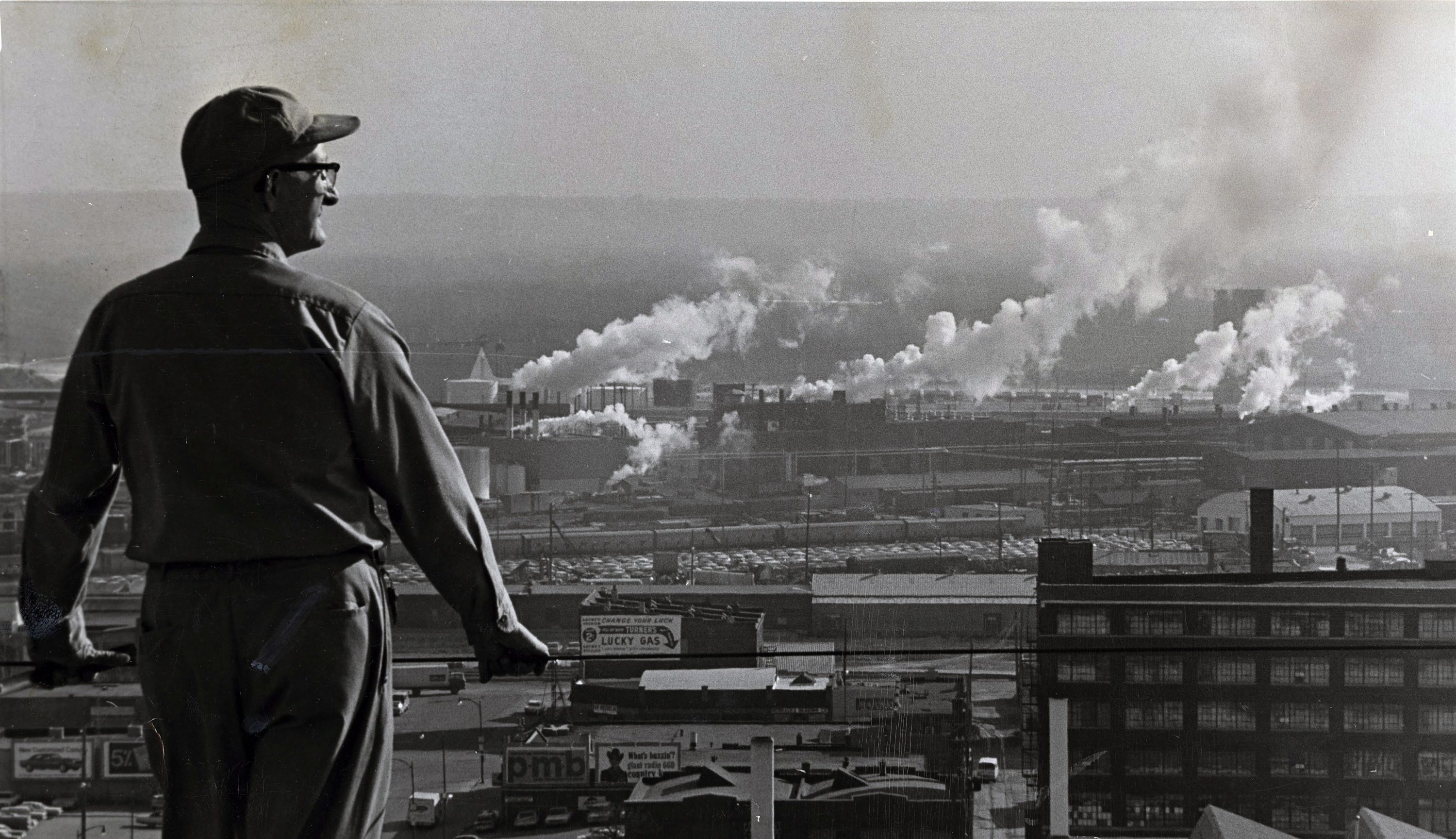 A black-and-white photo of a man leaning against a balcony and looking out onto a landscape of buildings and smog.