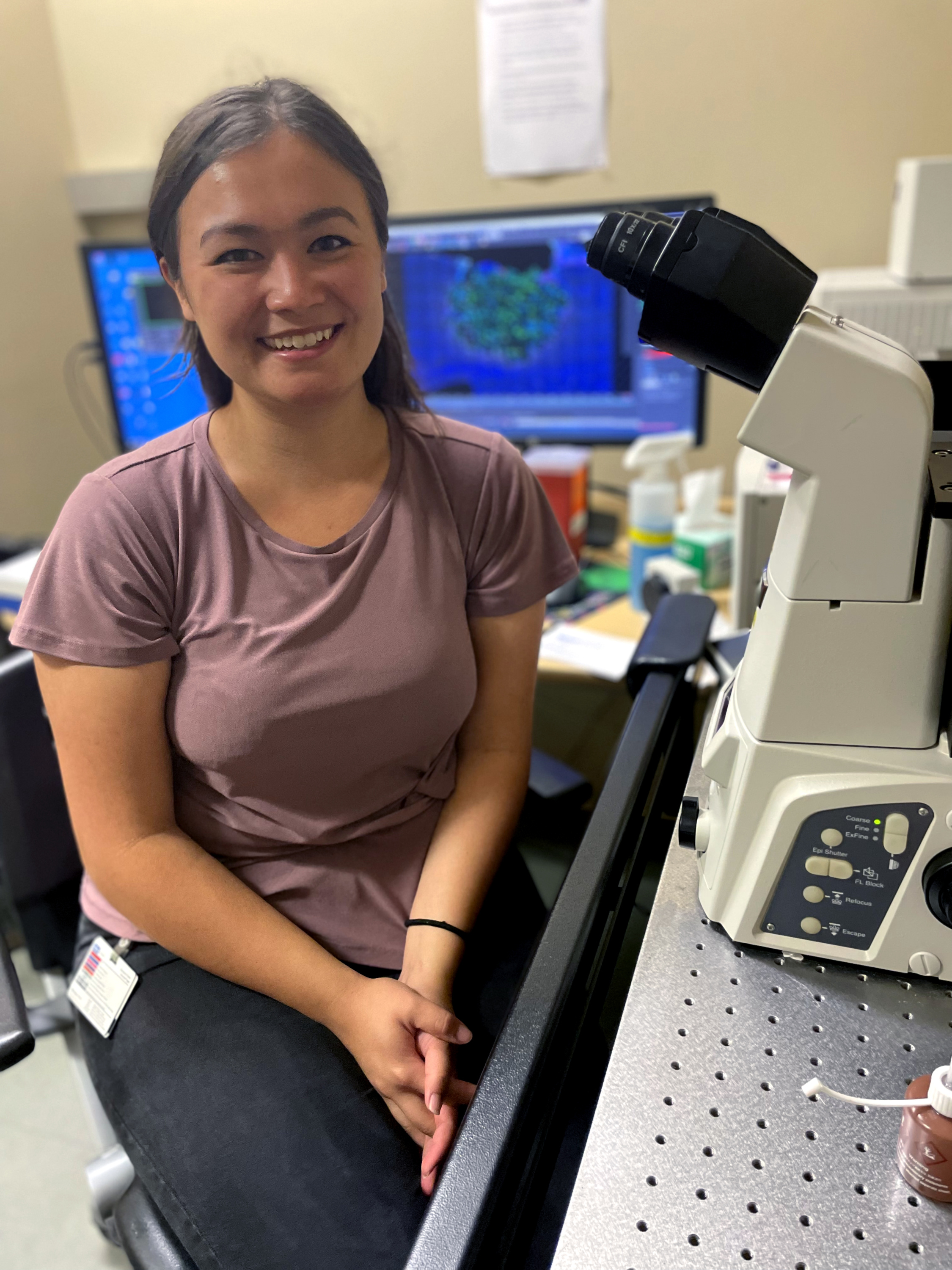 A woman wearing a pink T-shirt smiles and sits at a table with lab equipment.