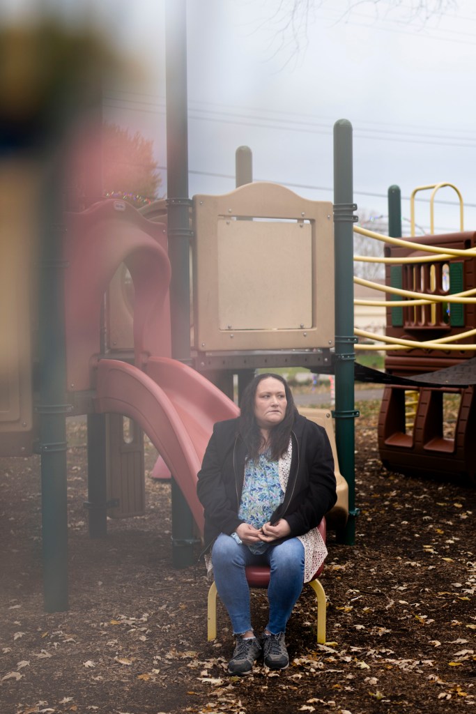 A woman with long brown hair, wearing a jacket and jeans, seated on a playground slide and looking off into the distance.