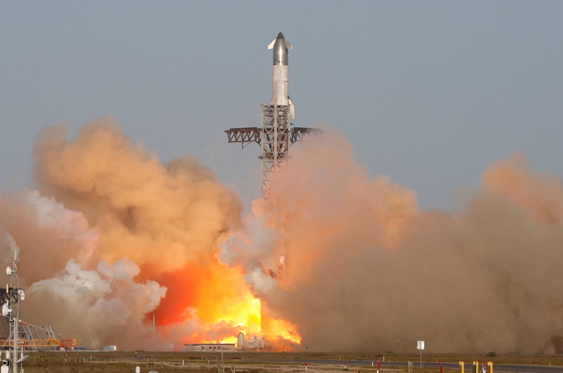 A large rocket launches into the sky next to a tall metal tower. The area around the tower is filled with smoke and fire.