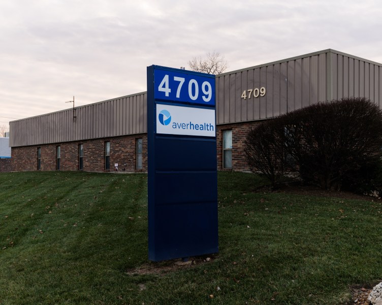 A tall, blue sign stands in neatly trimmed grass in front of a building with brown brick on the first floor and vertical brown siding on the second floor.
