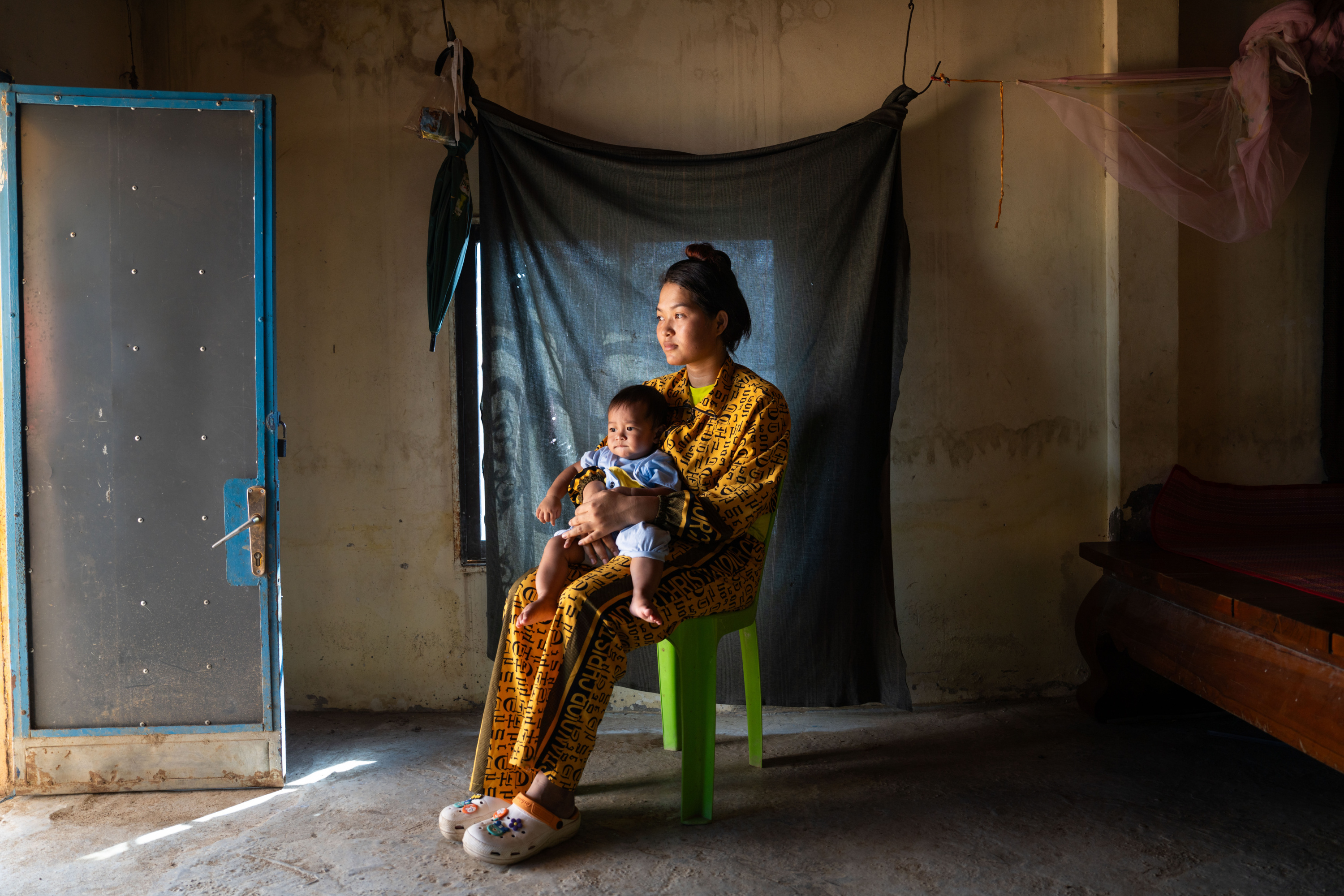 Eine Frau mit dunklem, hochgestecktem Haar hält ihren 8 Monate alten Sohn im Arm, während sie auf einem Plastikstuhl in einem Raum mit Betonboden sitzt.