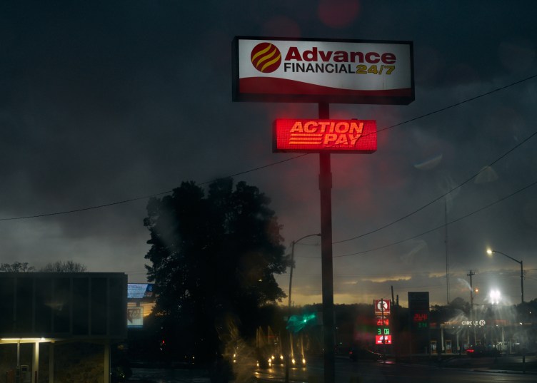 Two large signs on a tall pole rise above stores and streets. The top sign reads “Advance Financial 24/7,” and just below it the second sign reads “Action Pay.”