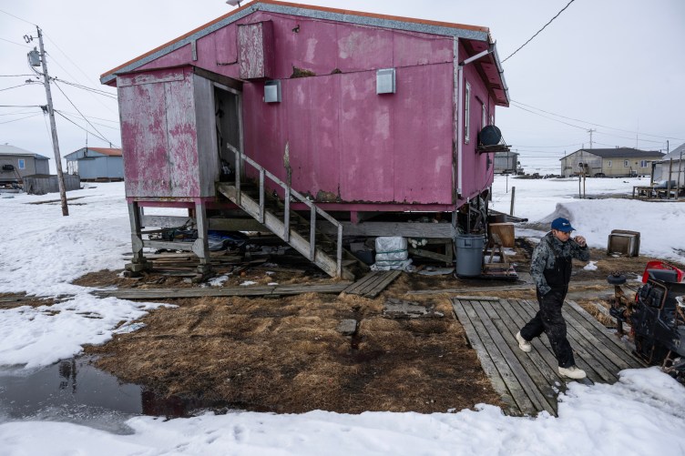 Un homme portant une combinaison et des bottes de travail et tenant une cigarette électronique marche le long d’une promenade en bois, loin d’une maison rose perchée sur de courtes échasses. Le sol est recouvert de neige.