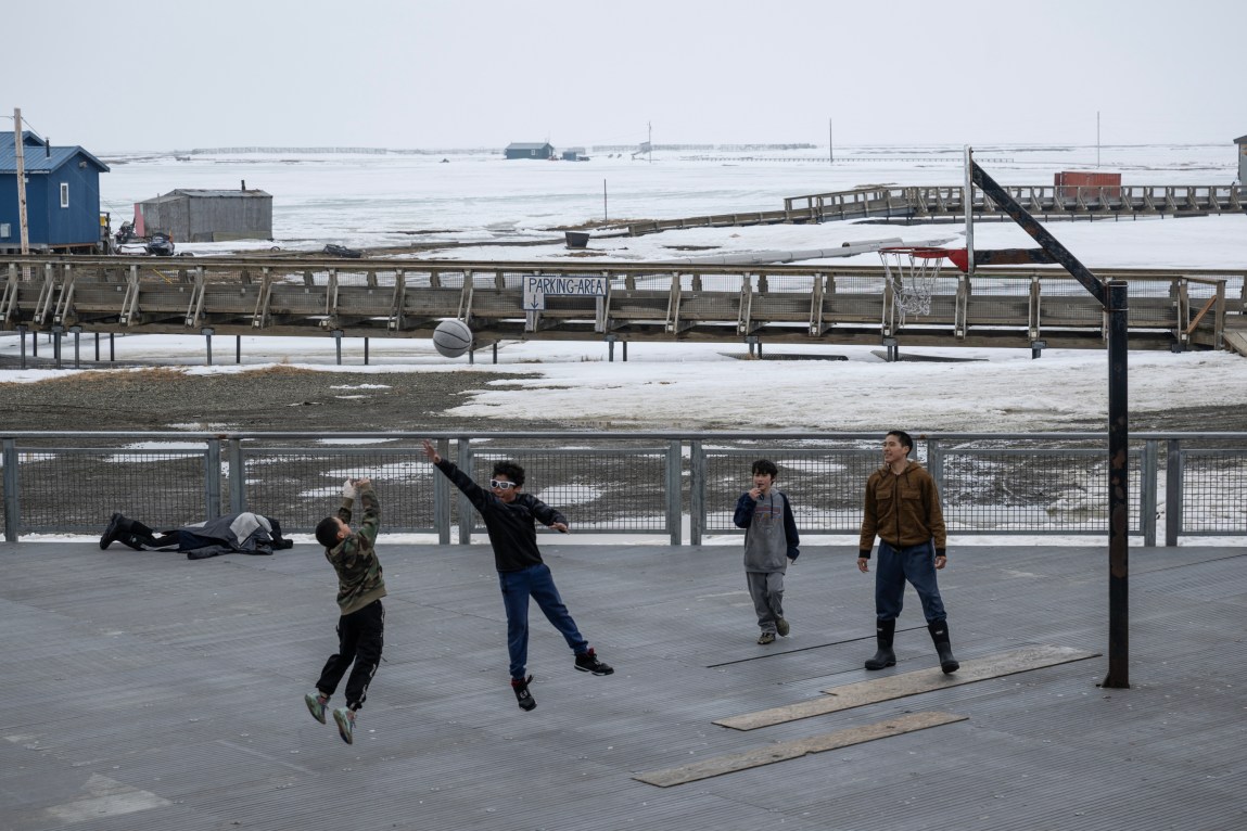 Deux enfants sautent en l’air pour jouer au ballon de basket gris pendant que deux autres se tiennent sur le terrain et regardent. Au-delà se trouvent un système de promenades grillagées et quelques bâtiments d'un étage dispersés sur un terrain enneigé.