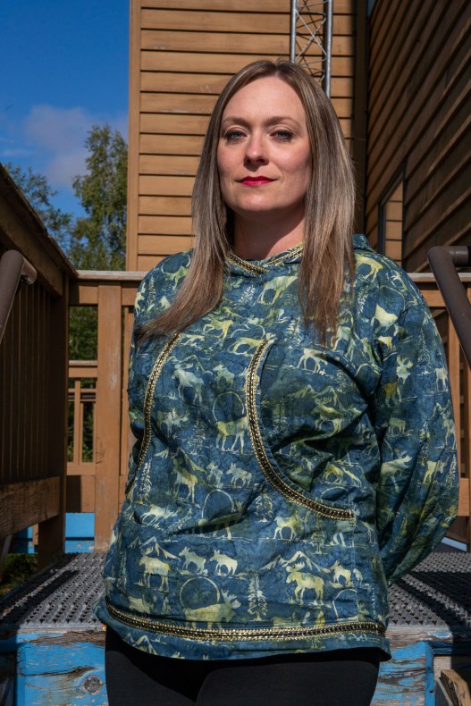 Portrait d'une femme debout sur un escalier extérieur portant une veste en tissu aux accents dorés ornée d'un motif d'orignal et de neige.