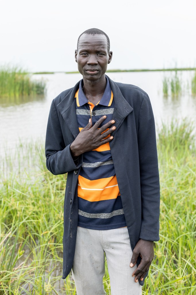 Ein Mann steht vor einer Fläche voller Wasser und Graspflanzen und hält seine rechte Hand über sein Herz.