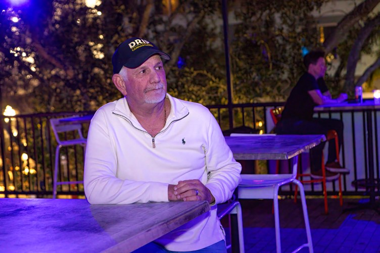 A man with white hair and a white mustache and goatee wearing a baseball hat and white sweater sits at a bar under indigo light.