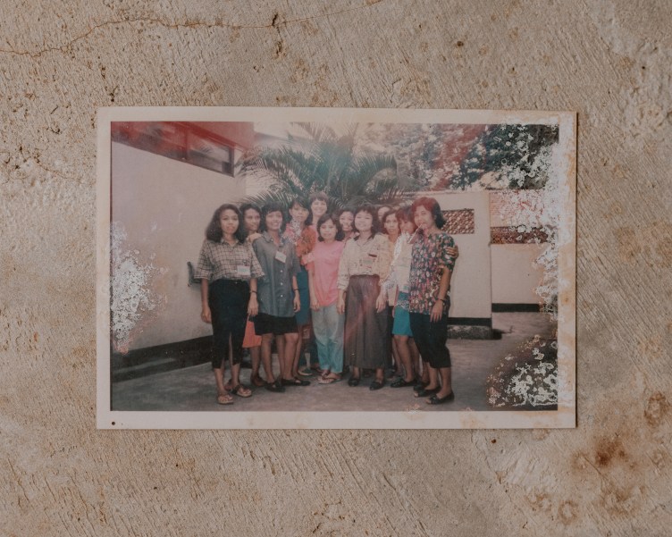 A faded photo with white damage on the edges shows a standing group of people wearing name tags and smiling. They are standing outside, in front of white walls, with plant fronds in the background.