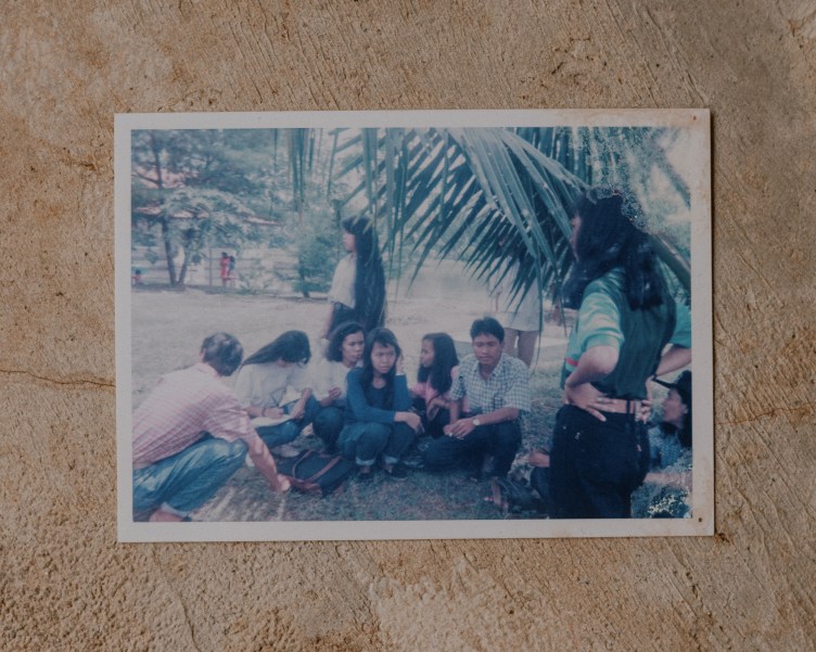 People sit in a circle outside on a lawn in the shade with trees in the background.
