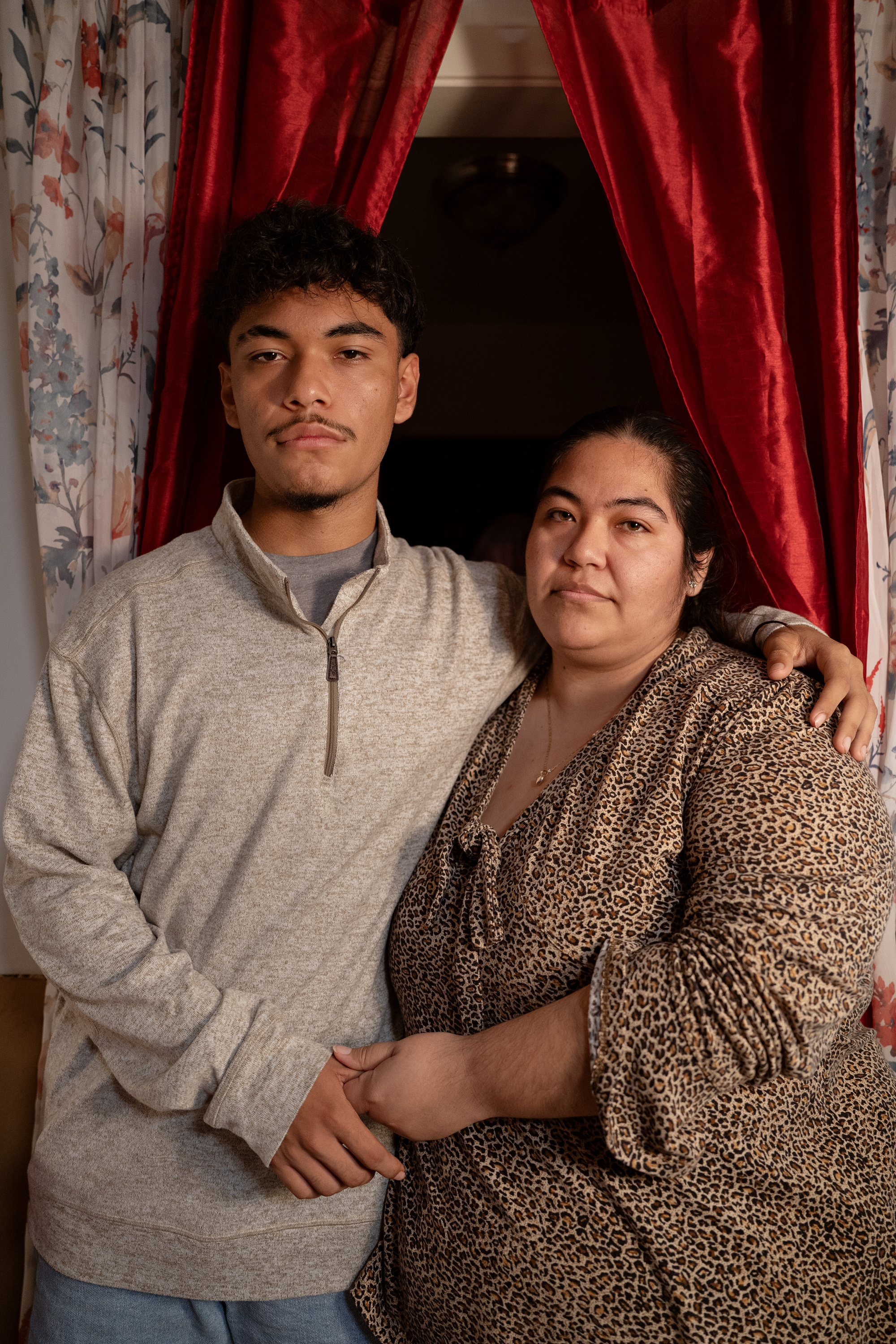 A young man with black curly hair and a thin goatee, wearing a gray long-sleeve shirt and blue jeans, poses for a picture alongside a woman with black hair and a gold locket around her neck, wearing a leopard-print shirt.
