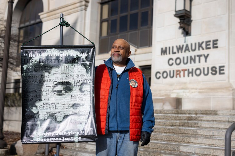 A bald man wearing blue-and-red winter clothing over a white T-shirt holds a sign with the photo of a young man while standing in front of the Milwaukee County Courthouse.