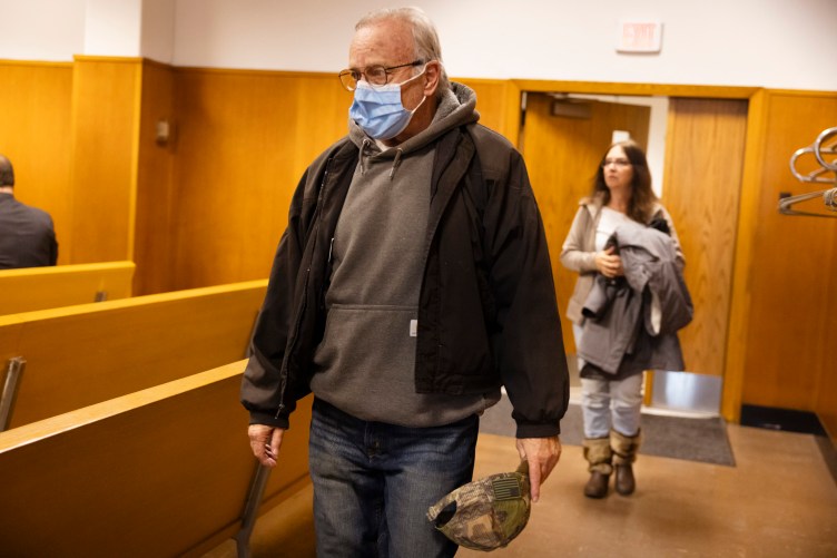 Un homme portant un masque, des lunettes et une veste sur un sweat-shirt entre dans une salle d’audience et porte une casquette de baseball camouflage. 