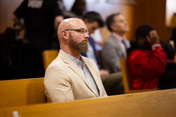 Un homme barbu et chauve, vêtu d'un blazer blanc, est assis dans une salle d'audience, entouré de personnes derrière lui. 
