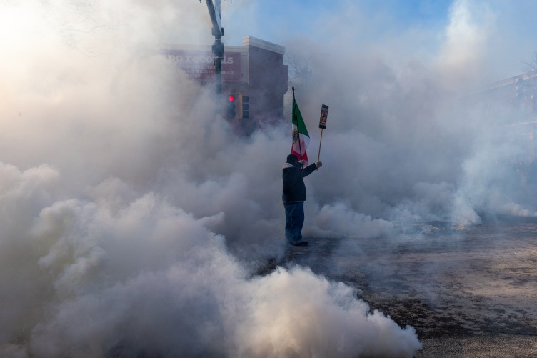 Ein Mann hält eine mexikanische Flagge und ein Protestschild in einer Tränengaswolke. Hinter ihm ist gerade noch eine Straßenlaterne zu erkennen, aber ansonsten ist die Gaswolke so dick und hoch, dass der Rest der Straße verdeckt ist.