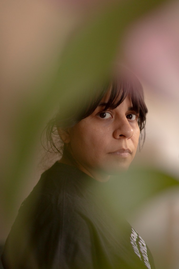 Une femme avec une frange brune et des cheveux tirés en arrière porte un T-shirt noir et de petites boucles d'oreilles. Elle regarde directement la caméra. Des formes floues roses et vertes les obscurcissent légèrement.