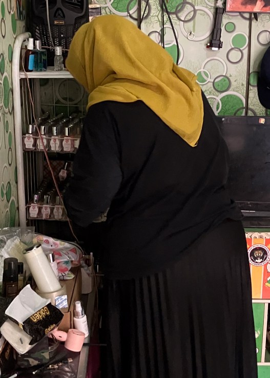 A woman wearing a yellow headscarf and black dress faces away from the camera toward the corner of a room filled with nail polish and other makeup. The walls are green and polka-dotted.